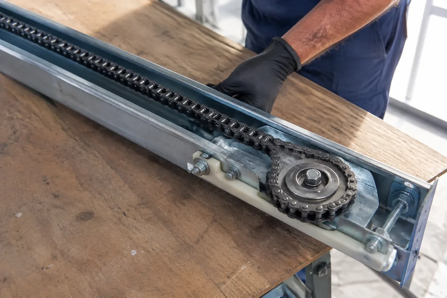 Gloved hand resting on a steel opener rail laid on a wooden bench, showing a drive chain looped around a sprocket wheel. — Chain-Drive Opener Repair, Richmond Hill